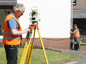 Bild vergrößern: Zwei Männer mit Warnweste vermessen mithilfe von Vermessungsgeräten ein Gebäude.