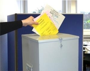 Bild vergrößern: Eine Hand wirft Wahlzettel in eine Wahlurne.