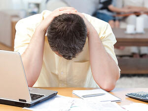 Bild vergrößern: Ein Mann sitzt gekrümmt mit beiden Händen hinter dem Kopf zusammengeschlagen mit Blick gerichtet auf die Schreibtischfläche.