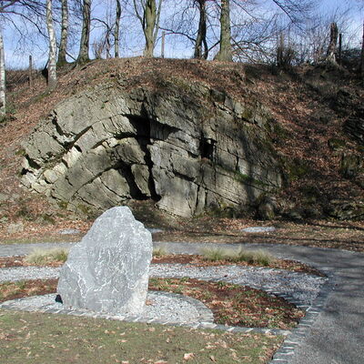 Bild vergrößern: Ein aufgestellter Stein vor einer Felswand geologischer Gesteinsschichten.