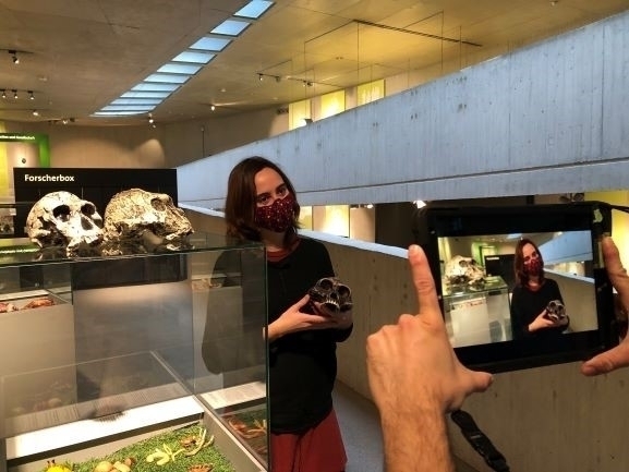 Bild vergrößern: Zwei Hnde fotografieren eine Schdel in der Hand haltende weibliche Person in einem Museumsaustellungsraum.