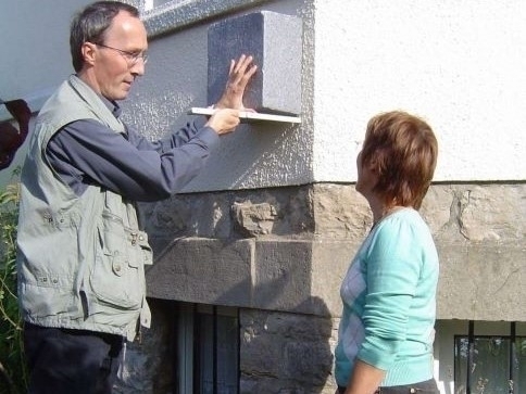 Bild vergrößern: Ein Mann misst mit einem Zollstock einen Stein an der Hauswand ab. Neben ihm steht eine Frau.